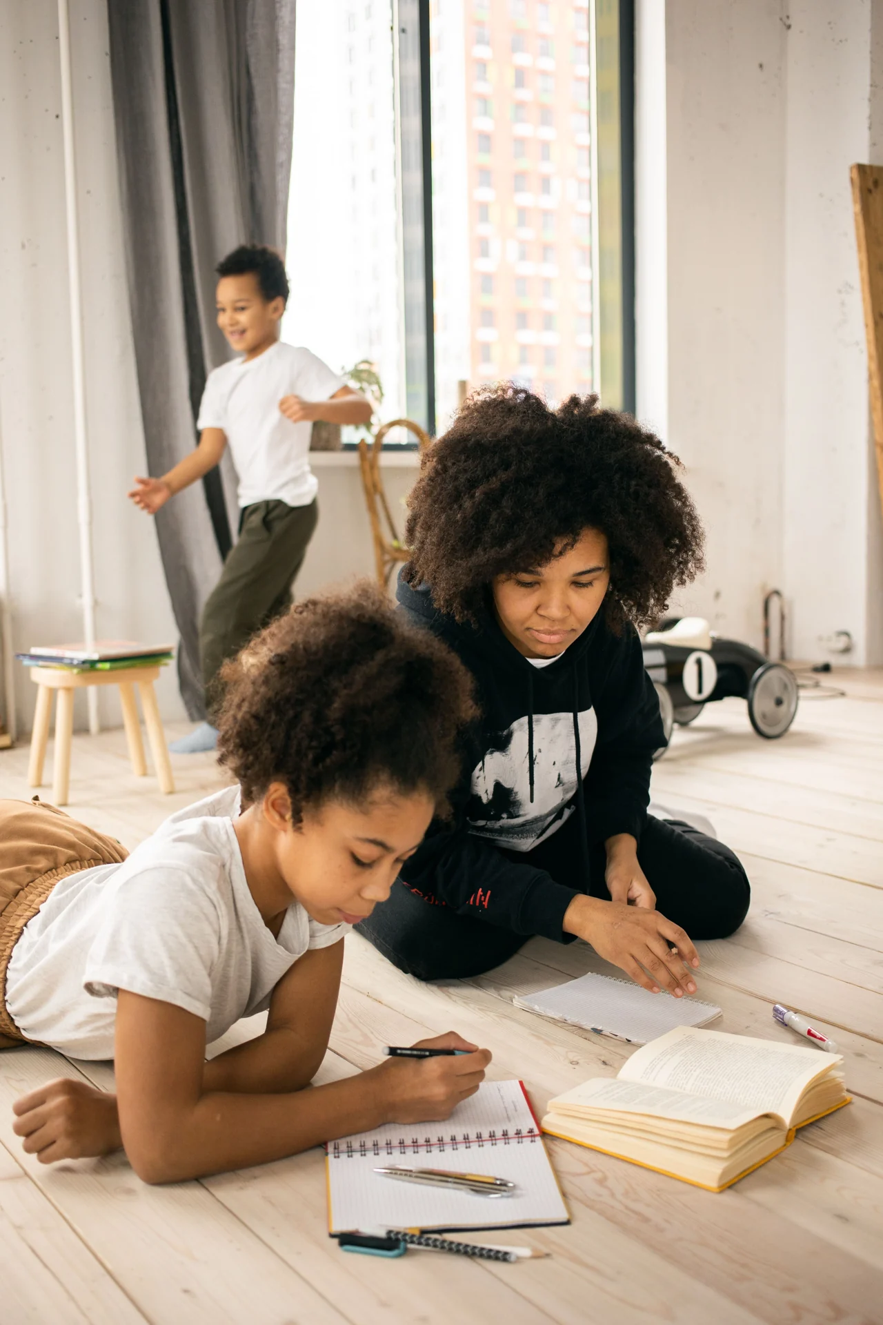 A mother helping her daughter with schoolwork at home, with her son playing nearby